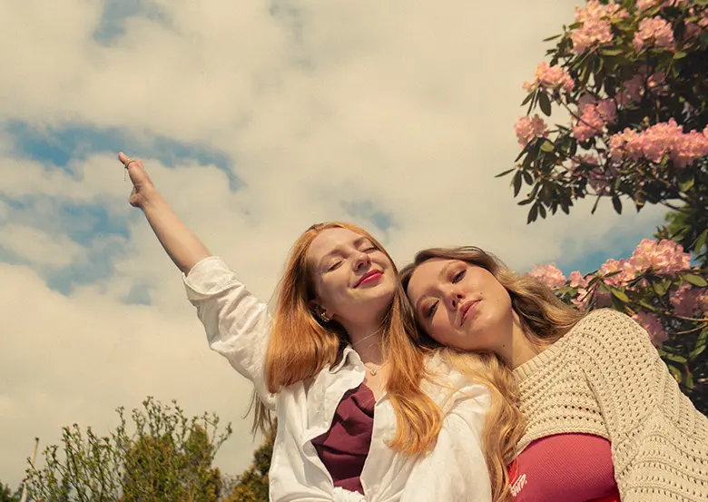 Two smiling young women under a blue sky beside a blossoming tree