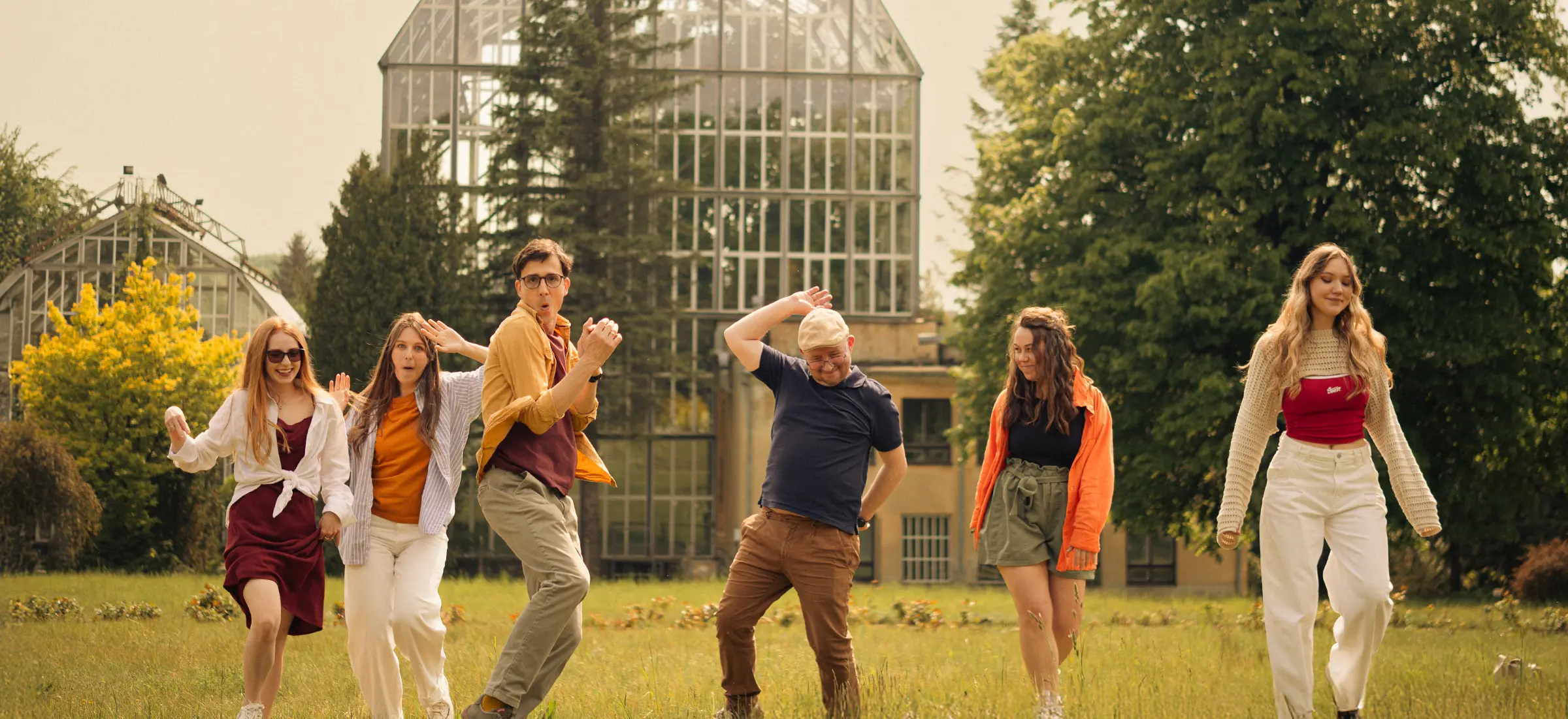 Group of people dancing joyfully on a lawn in front of a glass greenhouse